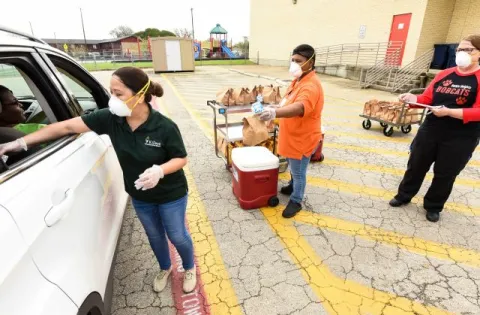 Hispanic woman with mask delivering food to vehicle. Two additional people in the background