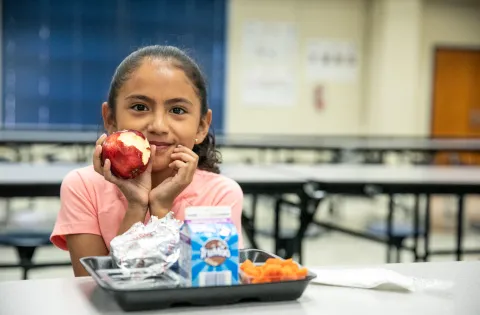 Latino girl eating apple in from of lunch tray