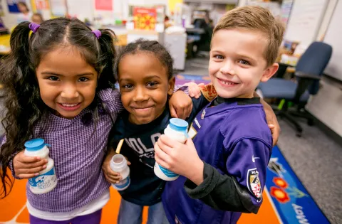 Three elementary school students--a Latina girl with pigtails, a Black boy with cornrows, and a white boy with blond hair--smiling while holding bottles of milk during school breakfast.