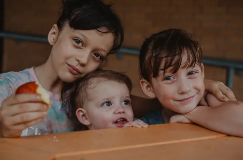 Three children leaning on table. One is holding an apple.